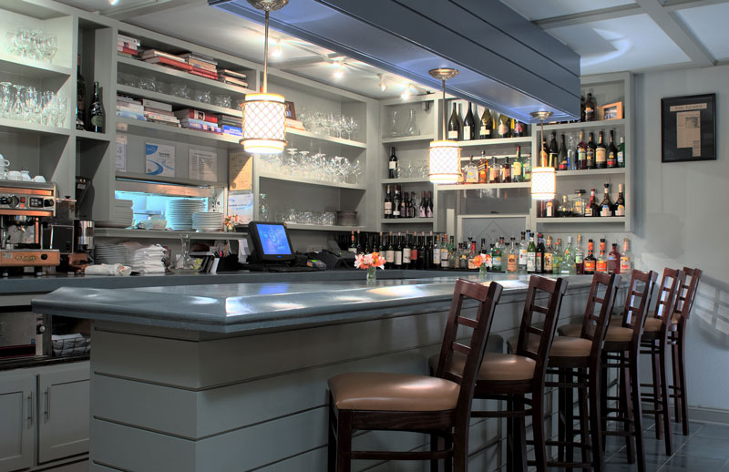 white and light blue bar counter and shelves lined with liquor bottles and glasses, and four chairs at bar